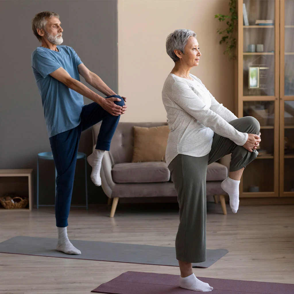 Two active older adults practising standing knee-hold balance stretches in a warm home setting